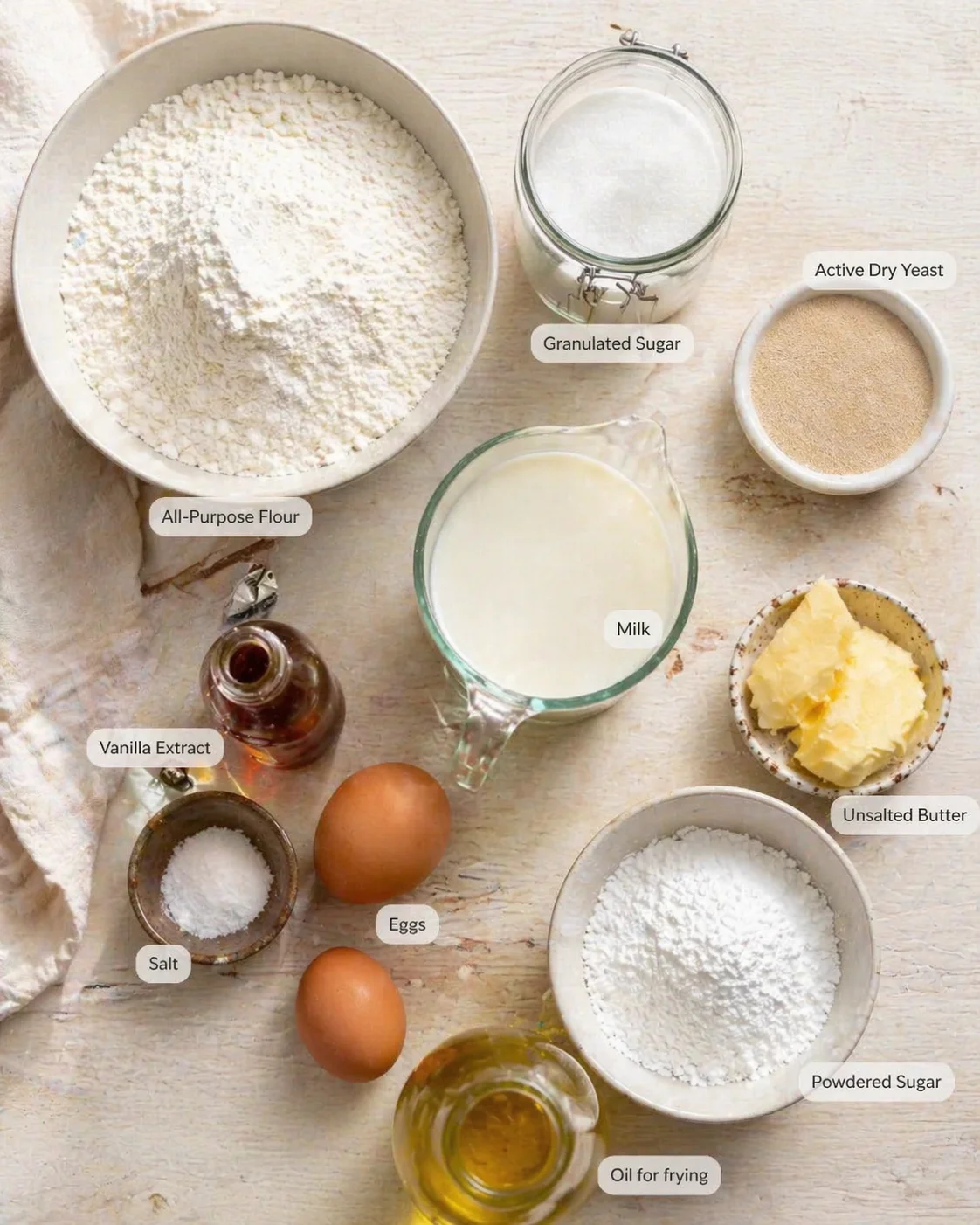 Top-down view of baking ingredients arranged on a light wooden surface, with flour, sugar, milk, eggs, butter, yeast.