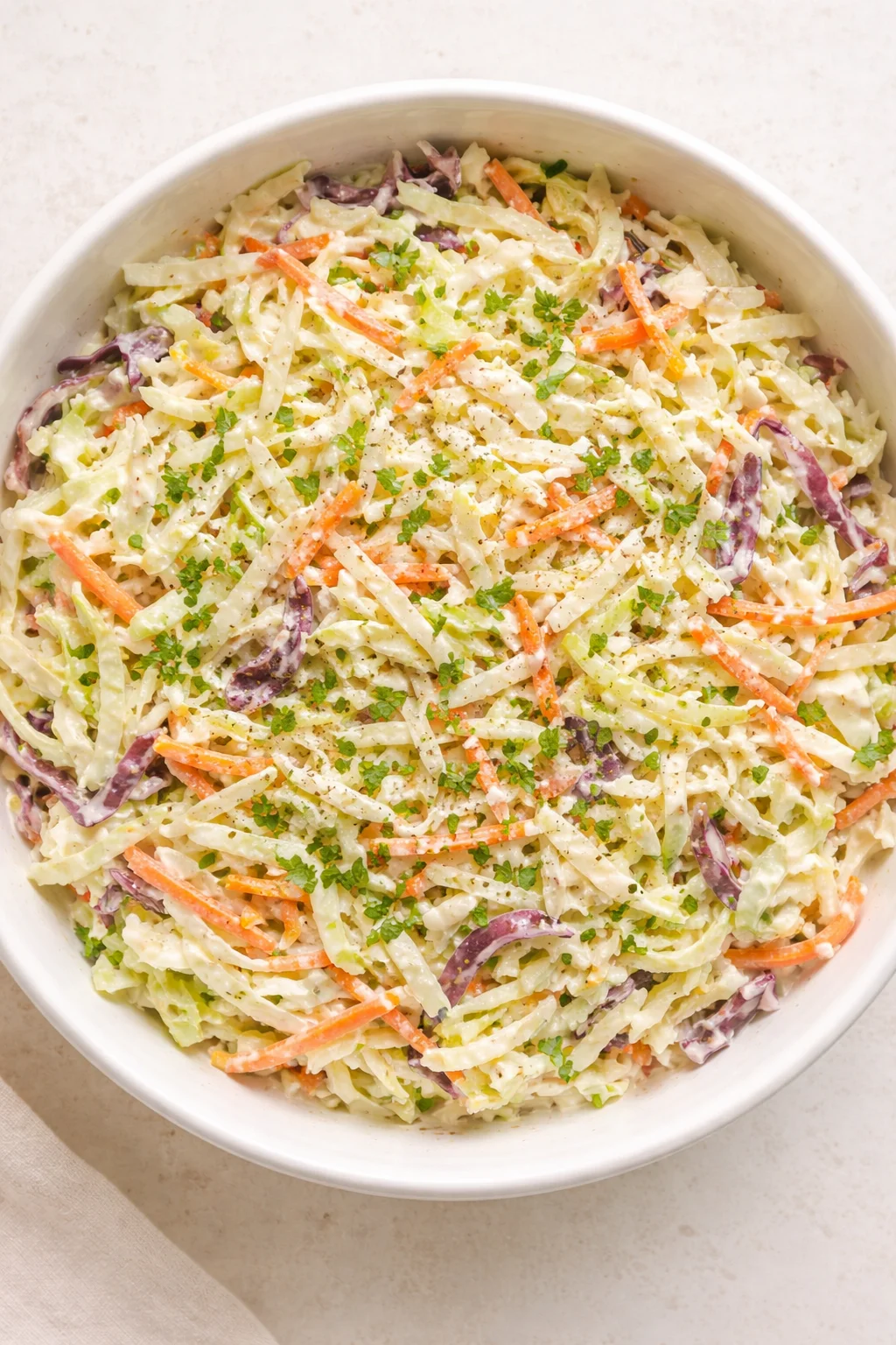 A white bowl filled with creamy coleslaw of shredded cabbage, carrots, purple cabbage, and parsley.