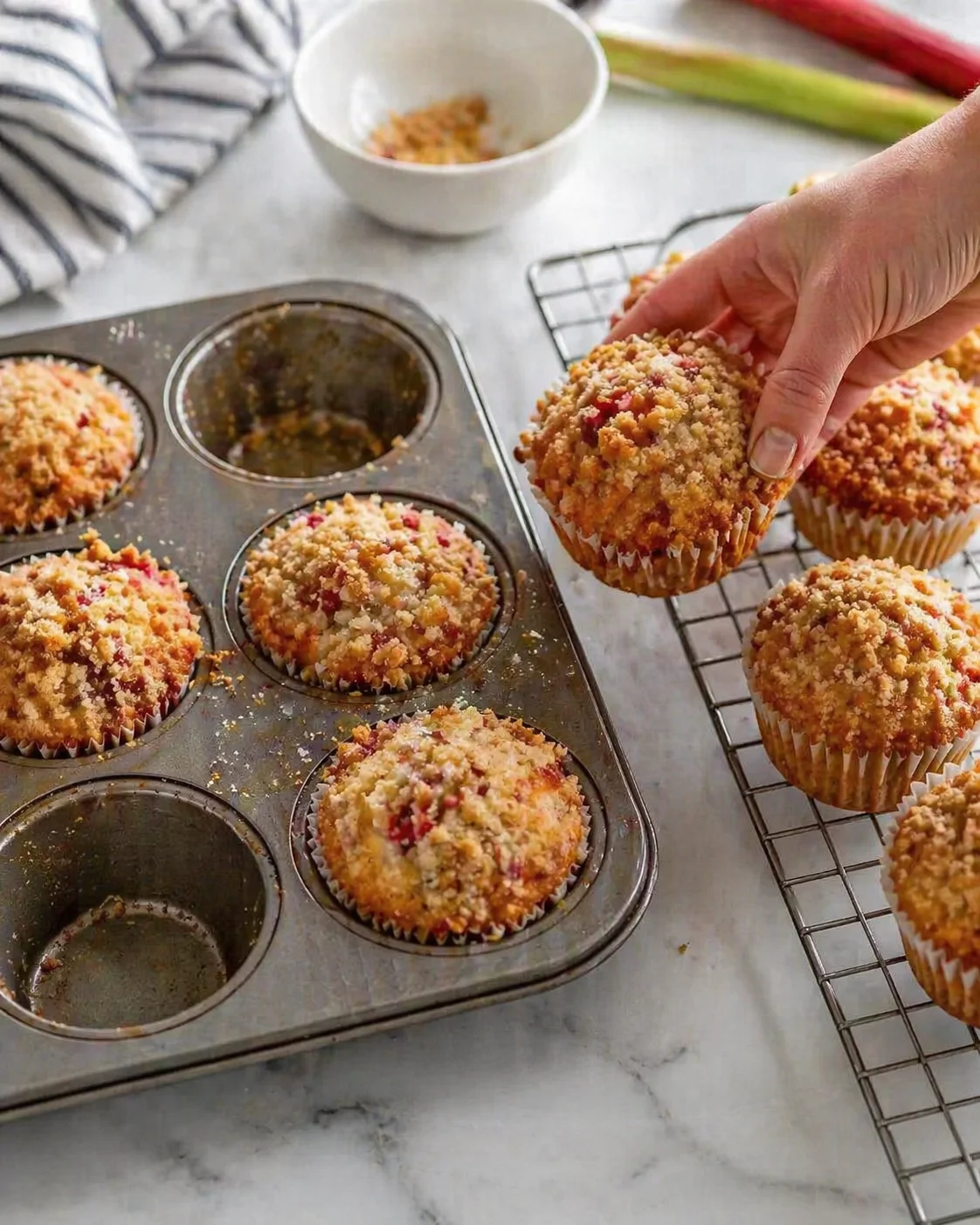 hand lifting a crumb-topped fruit muffin from a muffin tin onto a cooling rack