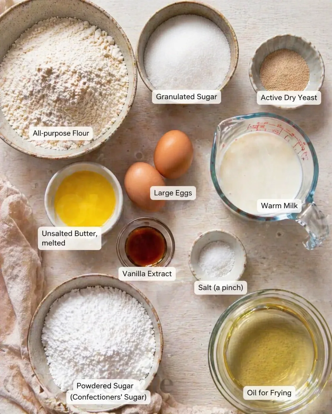 Overhead shot of labeled baking ingredients arranged on a light wooden surface, including flour, sugar, yeast, eggs, milk, and oil.
