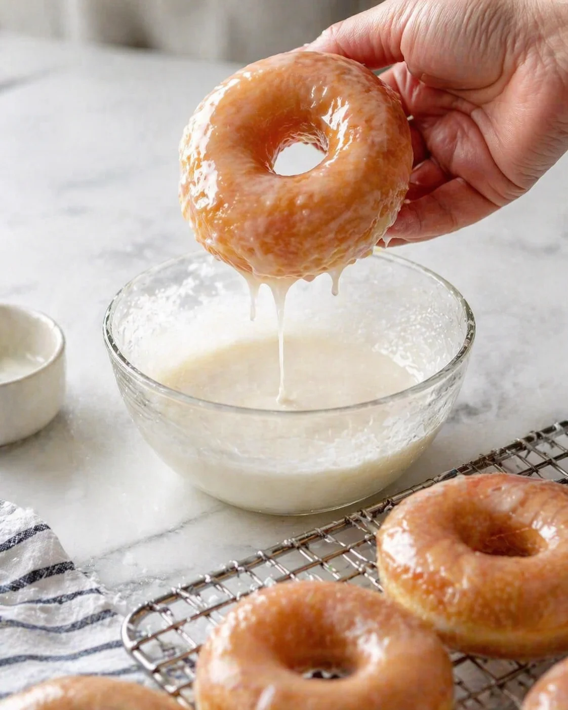 Close-up of a hand dipping a glazed donut into a glossy bowl of glaze.