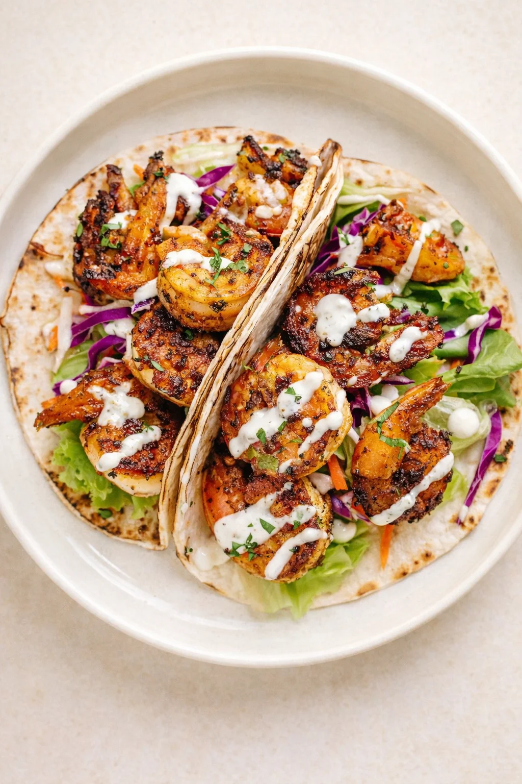 two tortillas hold charred shrimp atop greens with purple cabbage and cream drizzle on a light plate