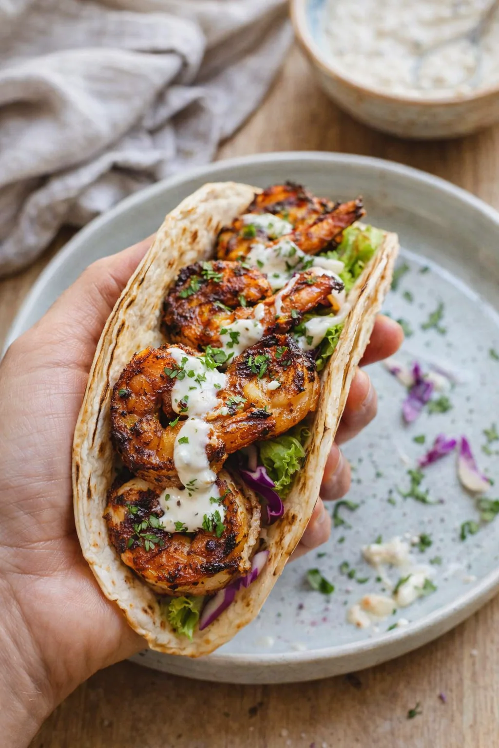 hand holding a flatbread wrap filled with grilled shrimp, greens, and a drizzle of creamy white sauce