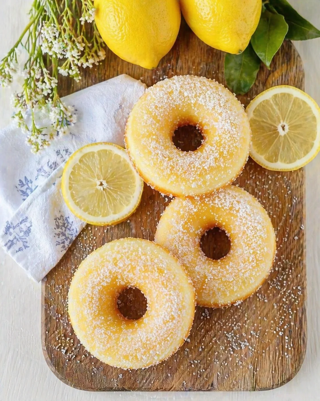 Three sugar-dusted lemon donuts on a wooden board with lemon halves and greens.
