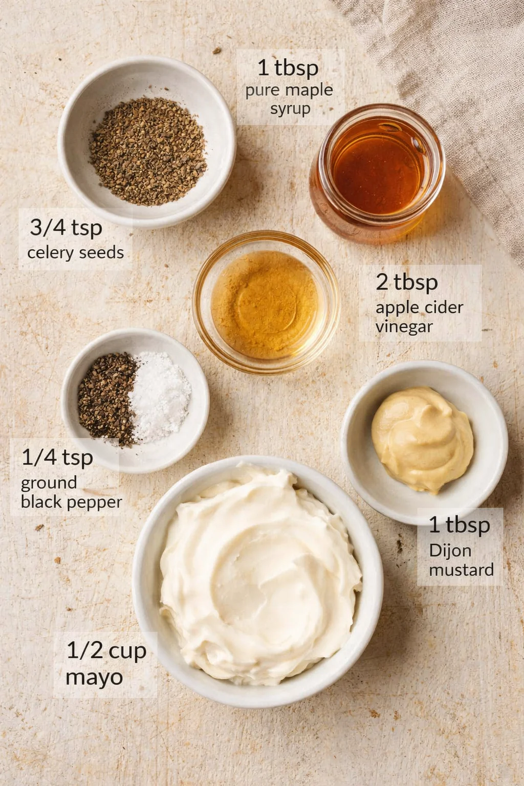 Overhead view of mayo, Dijon mustard, maple syrup, vinegar and spices arranged in small bowls on a light wooden surface.