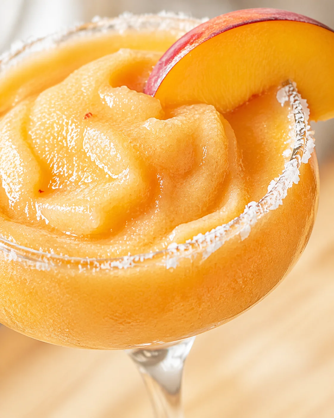 Bright orange peach-frozen cocktail in a salted-rim coupe glass with a peach slice garnish.