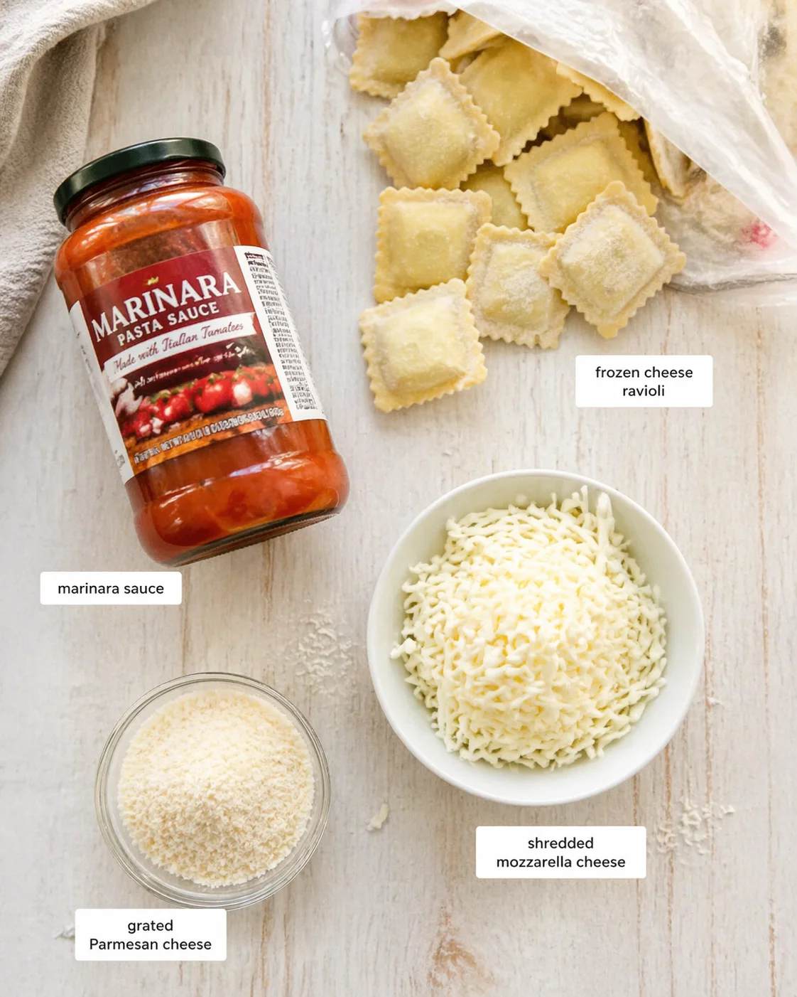 Top-down view of a marinara jar, frozen cheese ravioli, grated parmesan, and shredded mozzarella on a light wooden surface.