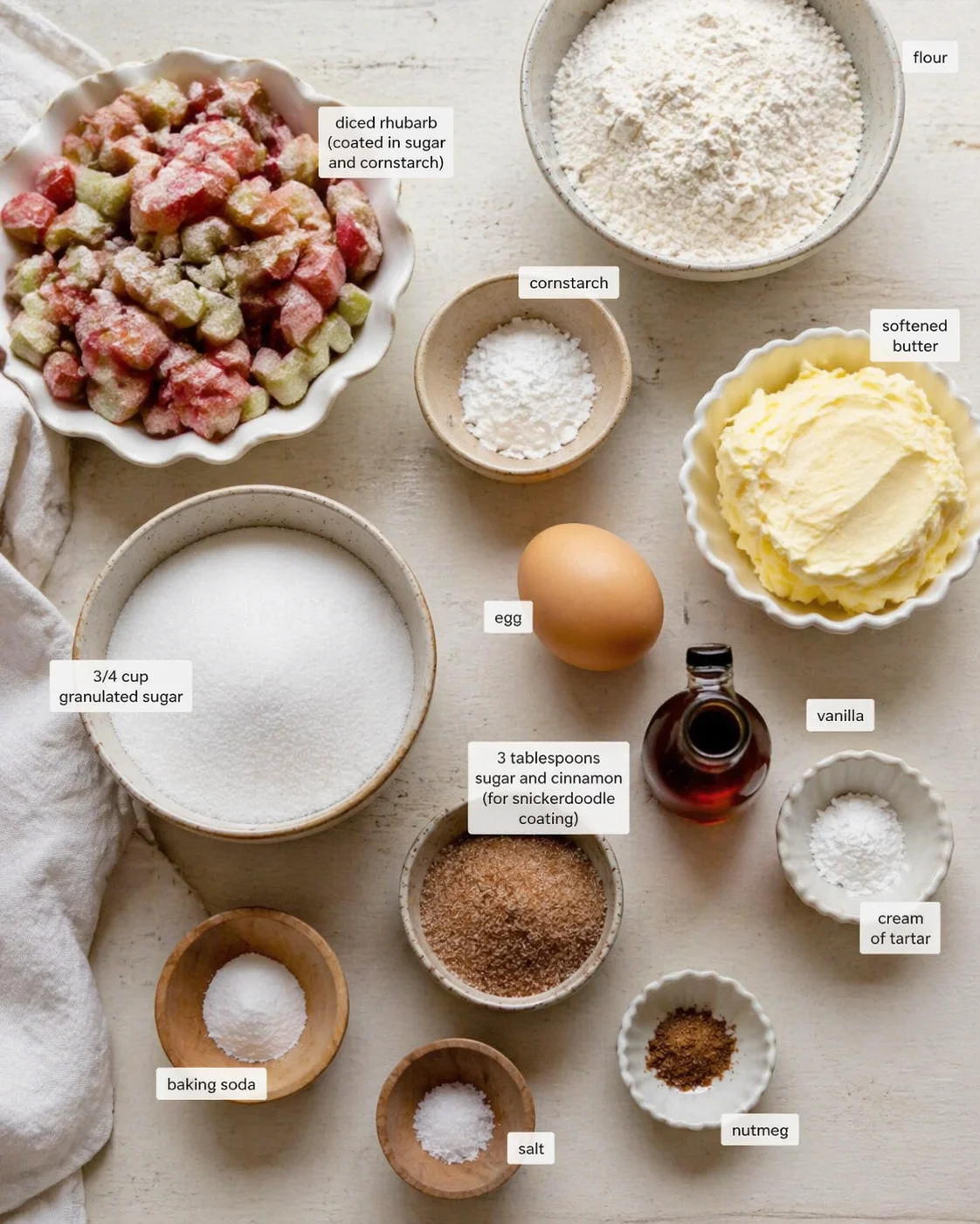 Top-down view of a baking ingredients spread with labeled bowls of rhubarb, flour, sugar, butter, egg, and spices.