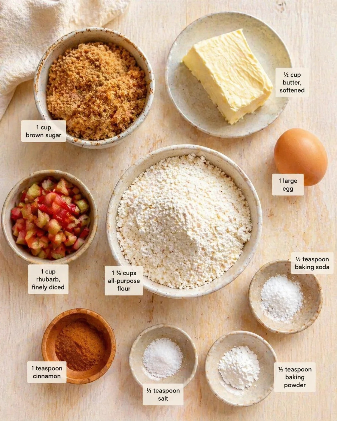Top-down view of rhubarb crumble ingredients arranged on a light wooden surface with labeled amounts.