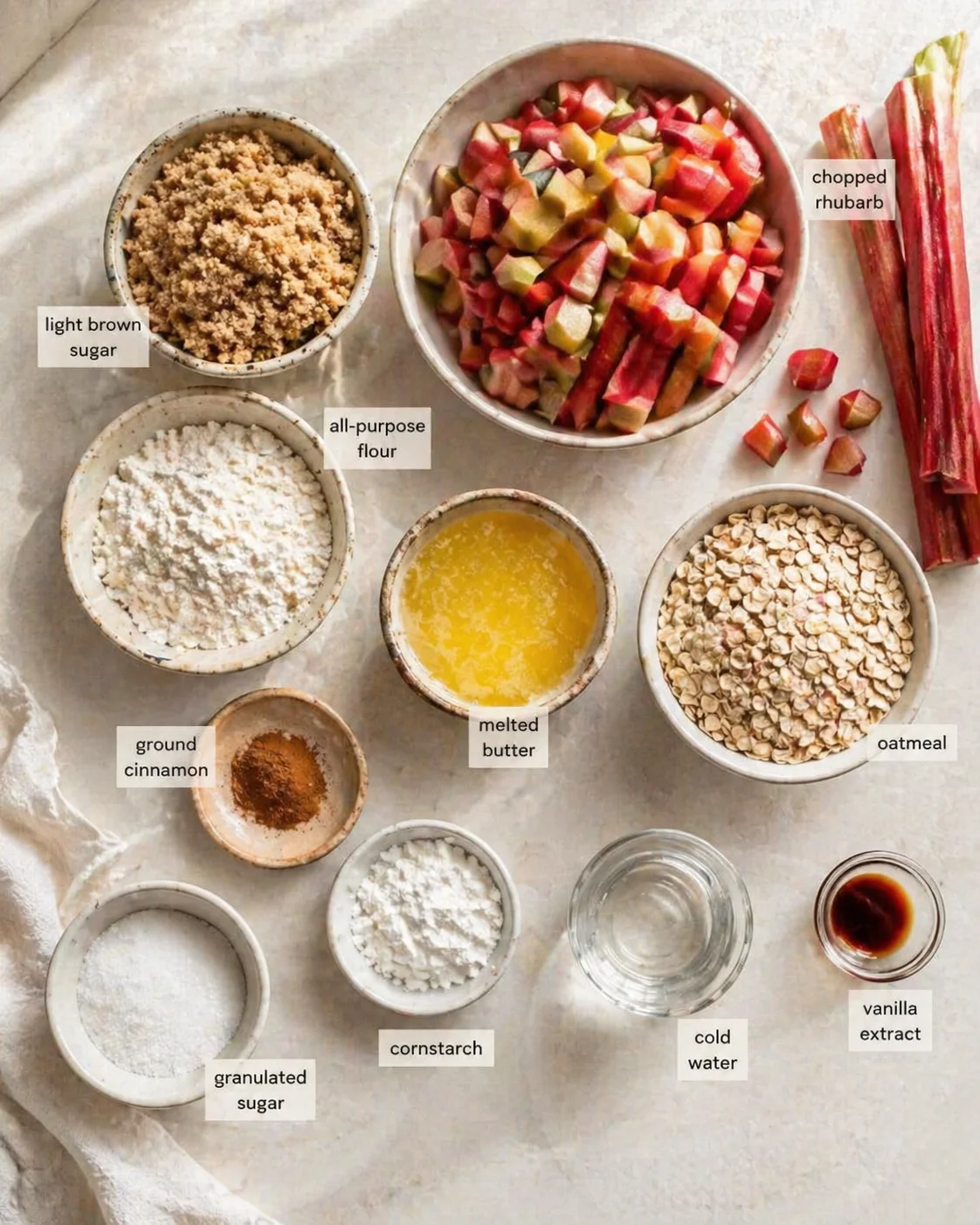 Top-down view of bowls containing rhubarb, oats, flour, sugar, melted butter, and spices on a light countertop.