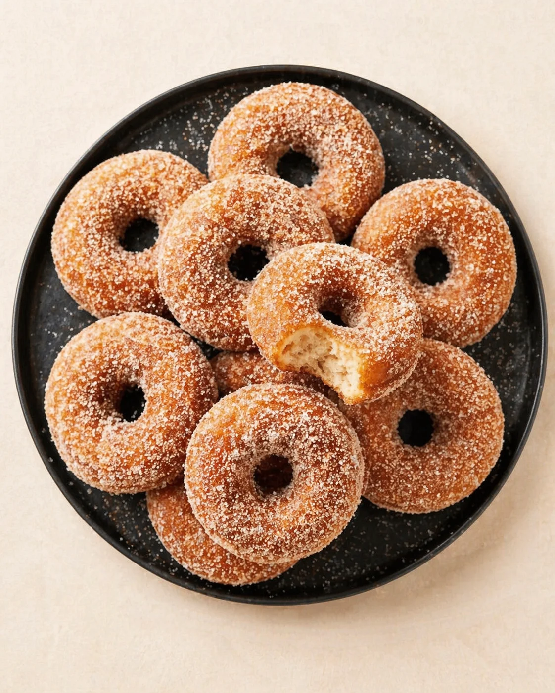top-down view of a black plate stacked with sugar-dusted ring donuts, with one broken bite revealing fluffy interior