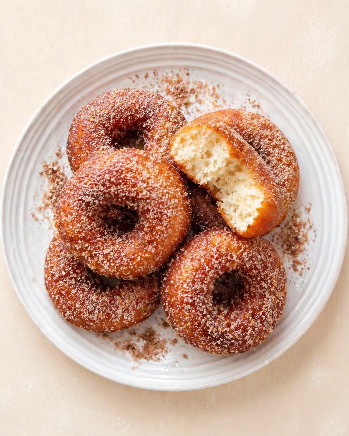 White ceramic plate filled with sugar-dusted donuts; a broken one reveals fluffy crumb and cinnamon dusting.