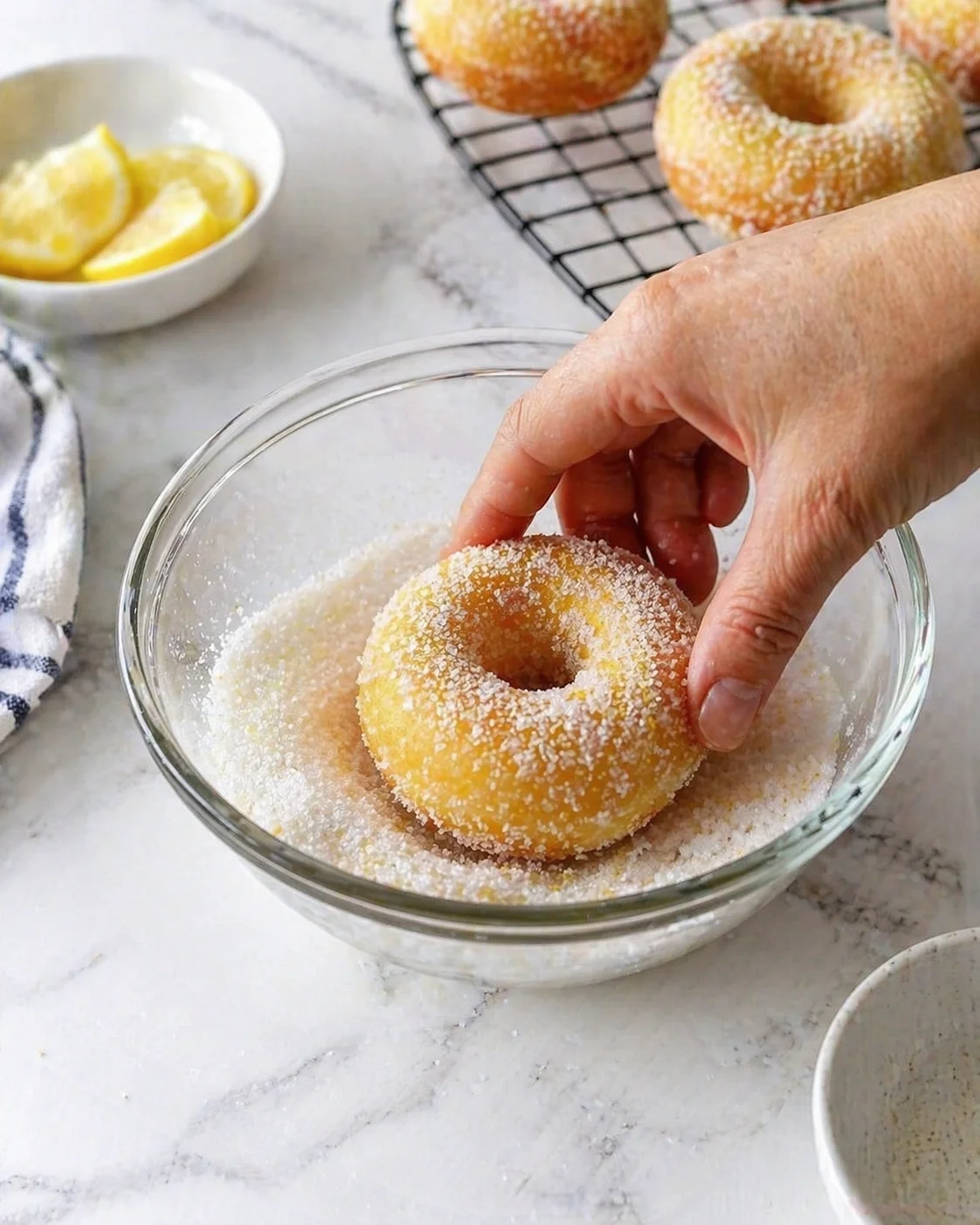 hand coating a golden sugar-dusted donut in a glass bowl on a marble counter