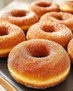 Close-up of golden, sugar-coated ring donuts arranged on a metal tray, warm lighting and textured crust.