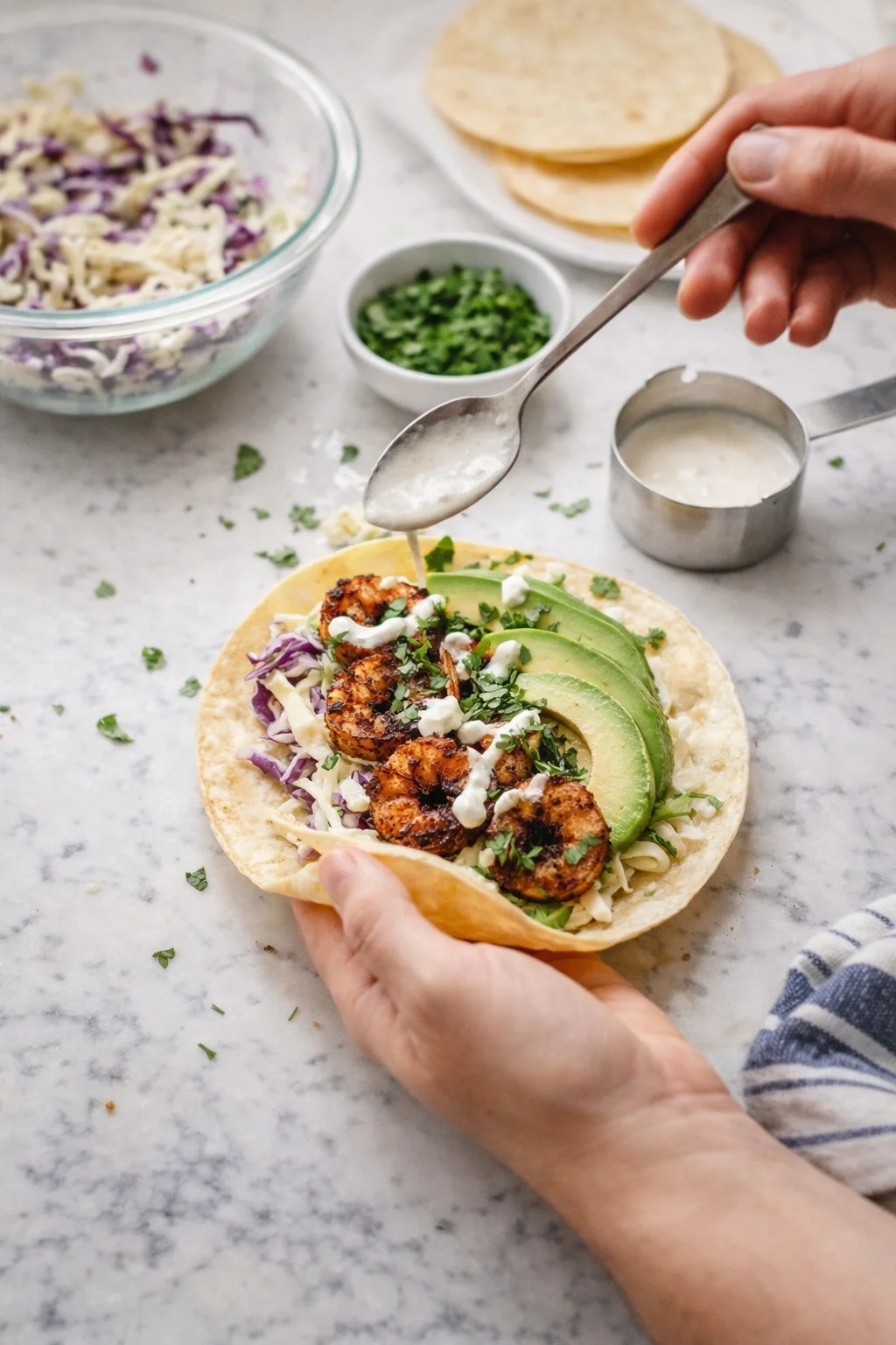 Close-up of a tortilla taco topped with avocado slices, cabbage slaw, and crema on a marble counter.