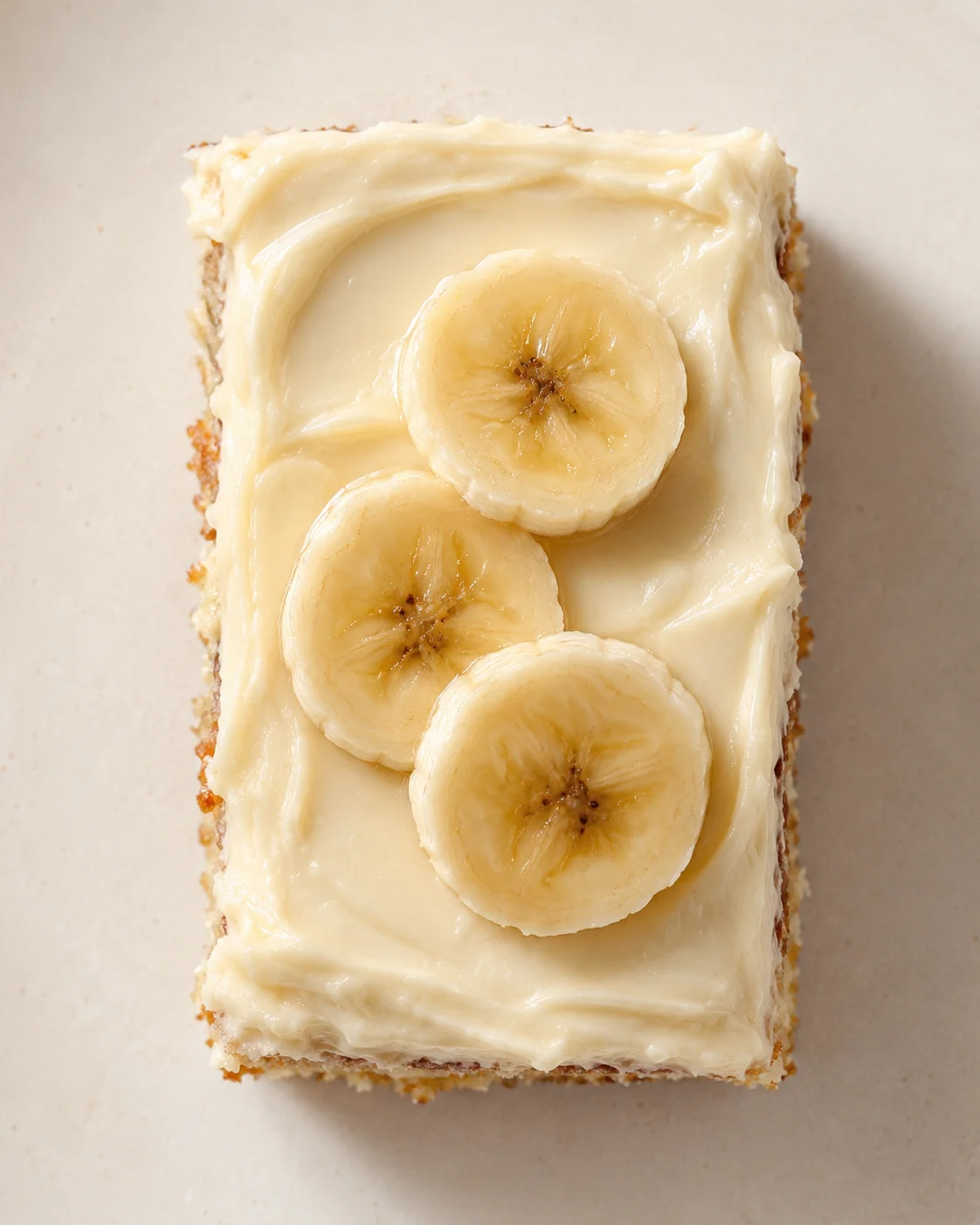 top-down view of a banana bread slice with cream cheese frosting and four banana rounds on a light beige background