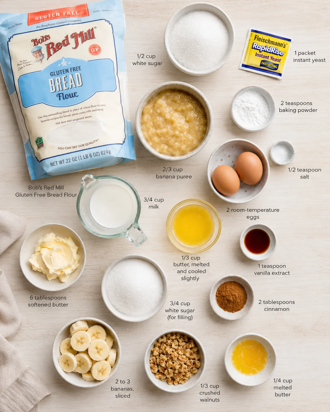 Overhead view of assorted banana bread ingredients arranged on a light wooden surface, including flour bag, eggs, milk, butter, sugar, bananas, walnuts, spices, and labeled measurements.