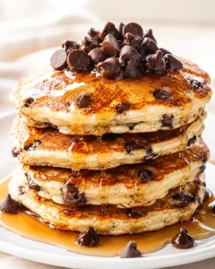 A towering stack of fluffy pancakes studded with dark chocolate chips, drenched in amber syrup on a white plate.