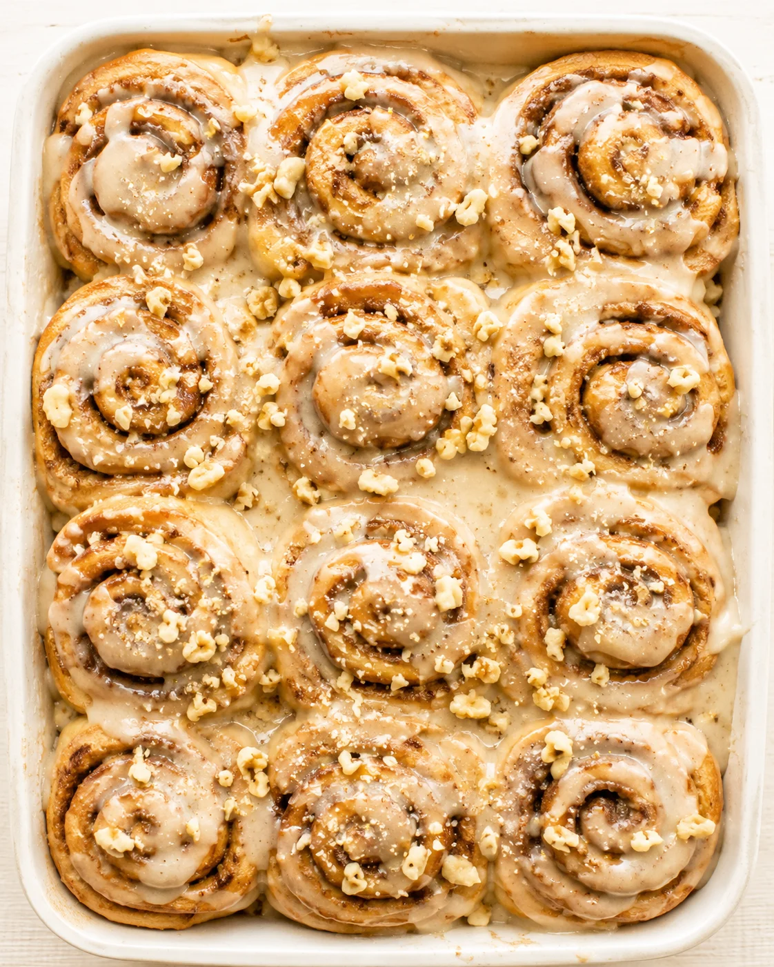 Top-down view of cinnamon rolls in a baking dish with creamy glaze and crushed nuts.