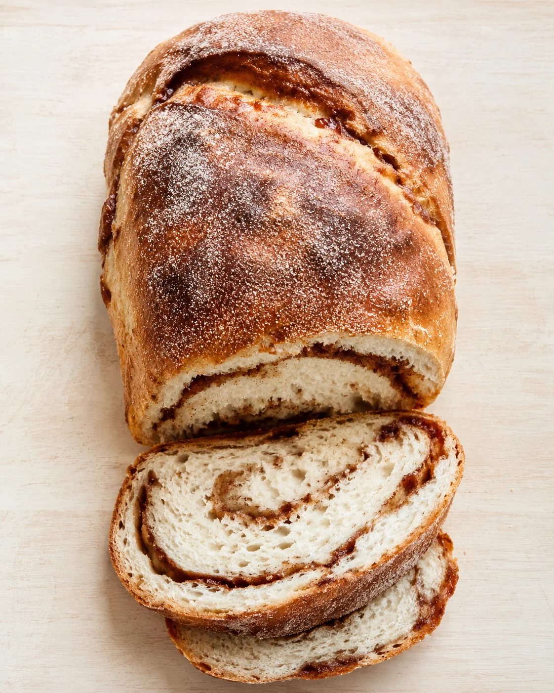 top-down view of a golden-brown cinnamon swirl bread loaf with flour dusting on a light wooden surface