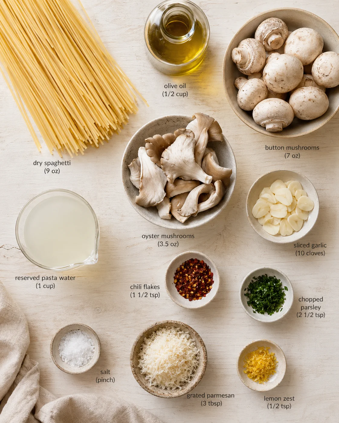 Top-down view of pasta ingredients arranged on a light wooden surface, including spaghetti, mushrooms, garlic, olive oil, parmesan, and herbs