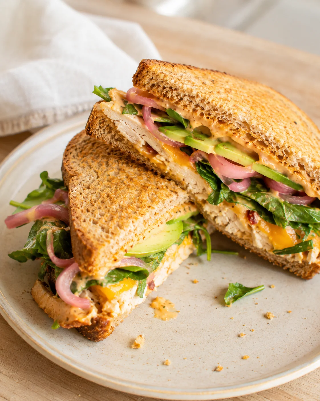 Close-up of a toasted multigrain chicken sandwich with avocado, pickled onions, greens, and creamy sauce on a beige plate.
