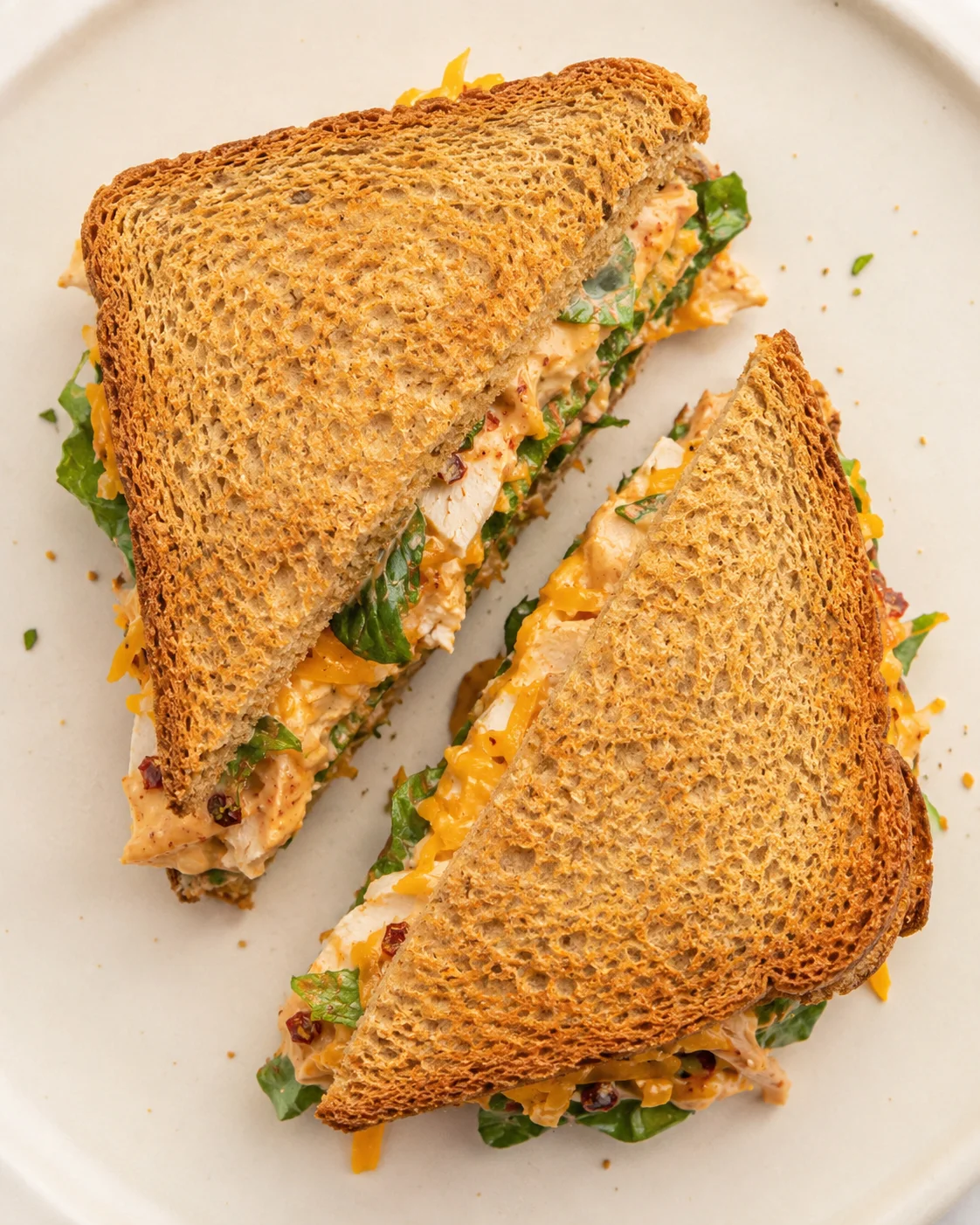 Two triangular toasted chicken-cheddar sandwiches on a pale plate, with greens peeking from the filling.