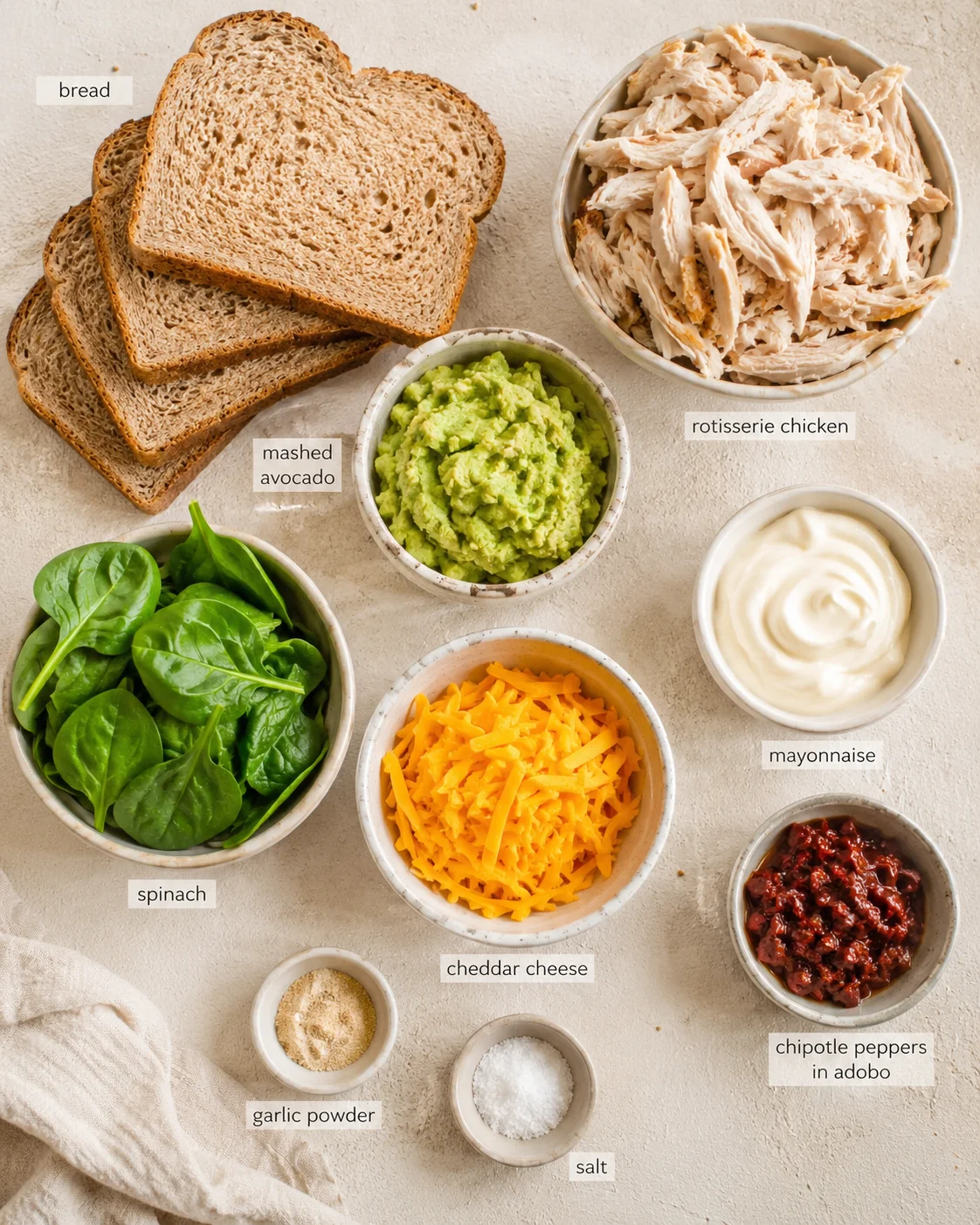 top-down flat lay of bread slices, rotisserie chicken, mashed avocado, spinach, cheddar cheese, mayonnaise, and chipotle peppers in adobo.