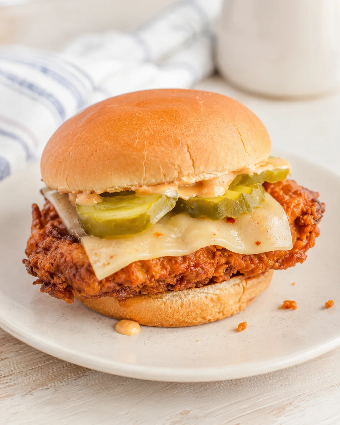 Golden fried chicken sandwich with pickles and melted cheese on a soft bun, on a white plate.