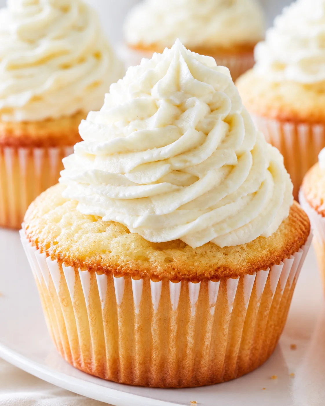 Close-up of a golden vanilla cupcake crowned with white buttercream frosting on a white plate.