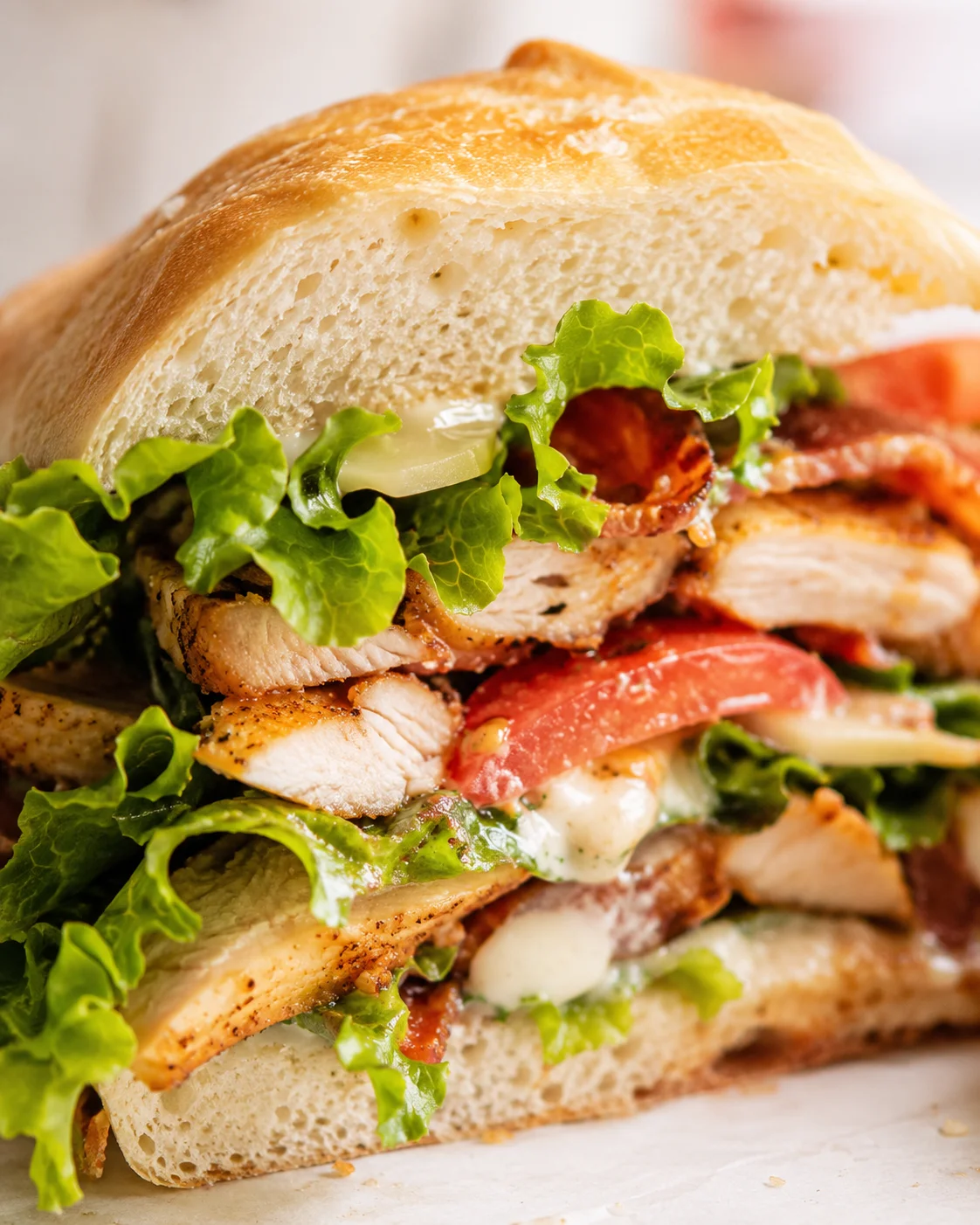 Close-up of a crusty artisan bun sandwich with grilled chicken, bacon, lettuce, tomato, and melted cheese.