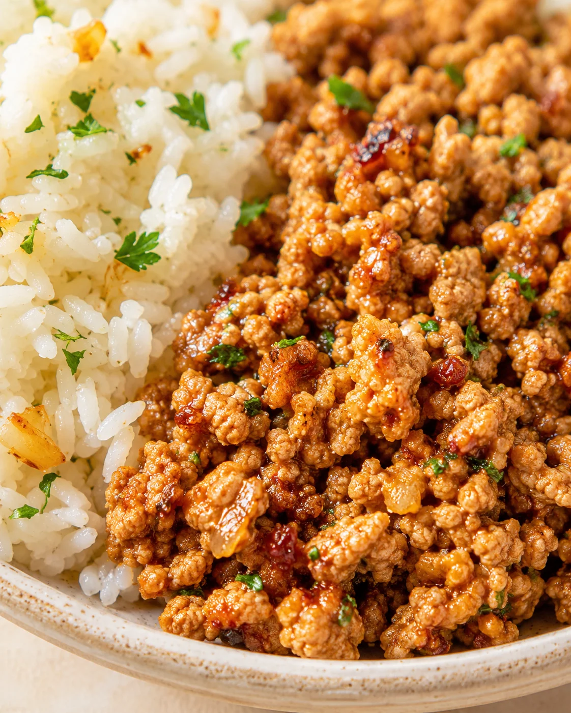Close-up of browned ground meat with herbs beside fluffy white rice in a beige bowl