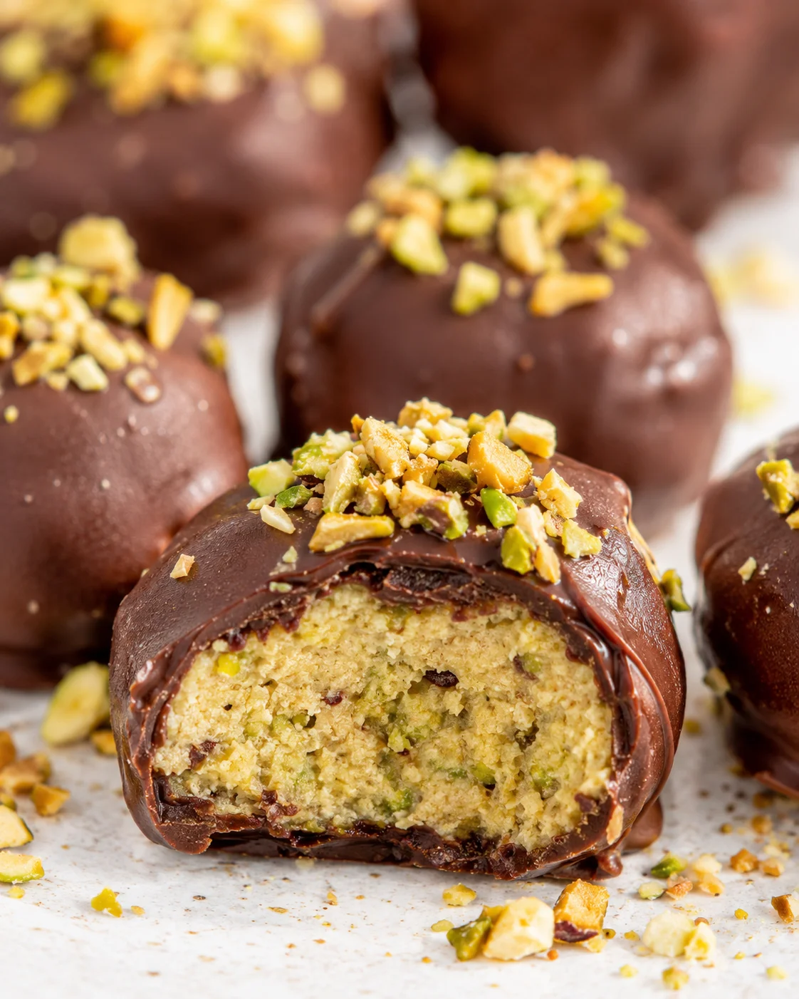 Close-up of chocolate-coated pistachio truffles topped with chopped pistachios, one bitten to reveal crumbly green-speckled interior.
