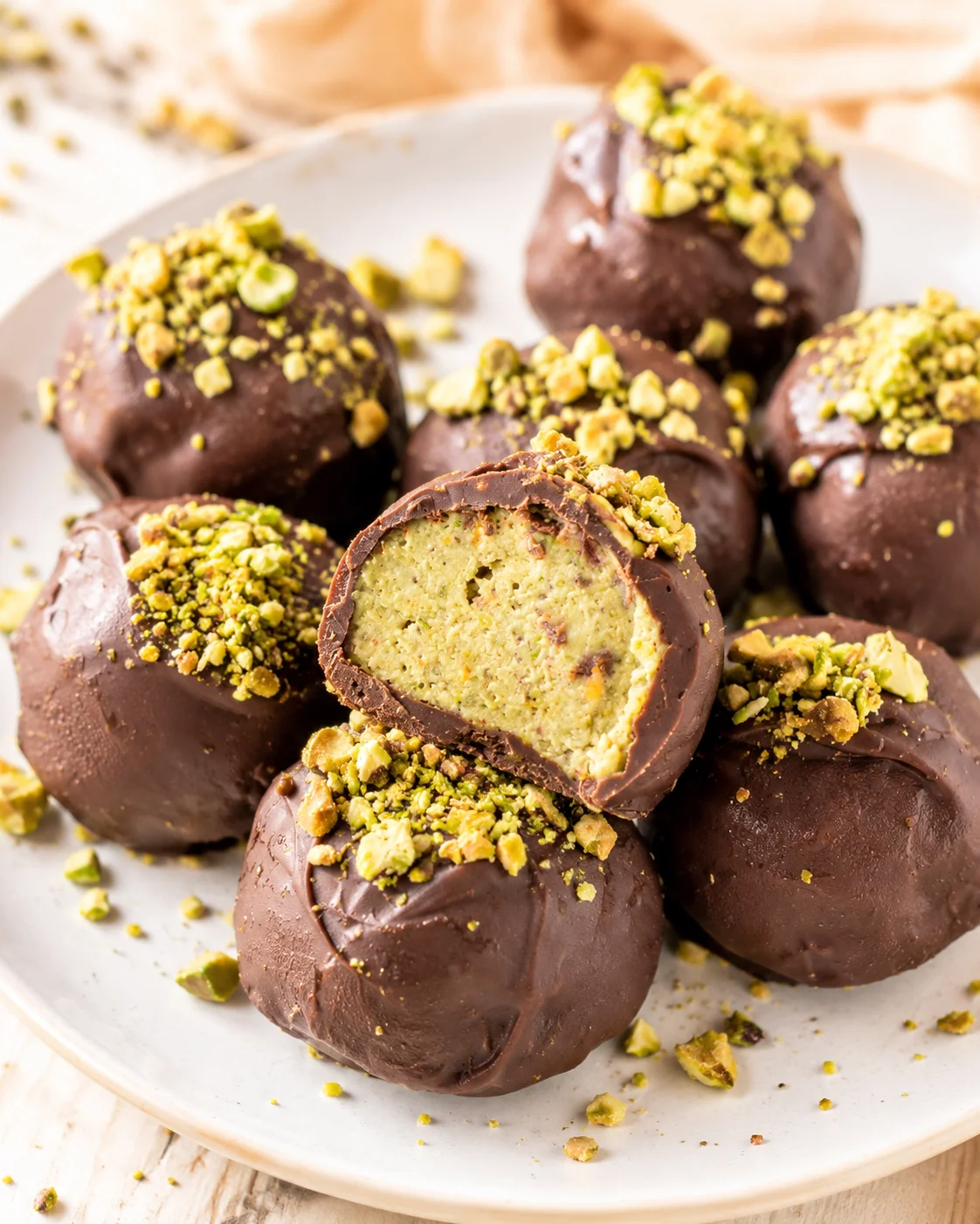 Chocolate-dipped pistachio truffles on a plate with chopped pistachios and one bitten to show green pistachio filling