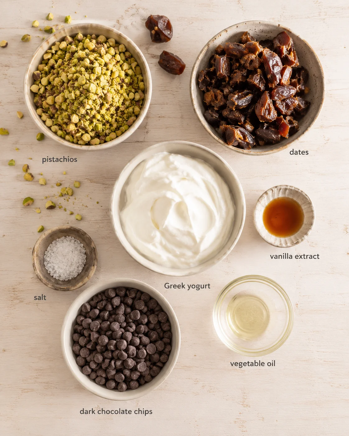 top-down view of assorted ingredients around yogurt: pistachios, dates, dark chocolate chips, vanilla extract, salt, oil