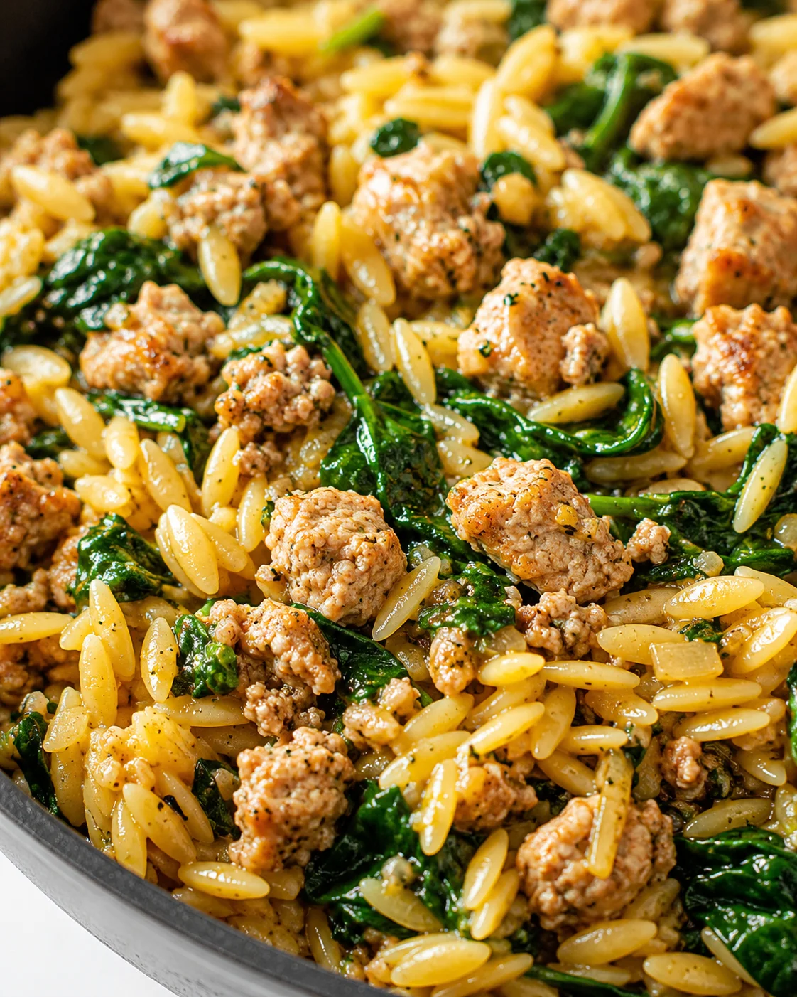Close-up of orzo pasta with sausage crumbles and wilted spinach in a skillet
