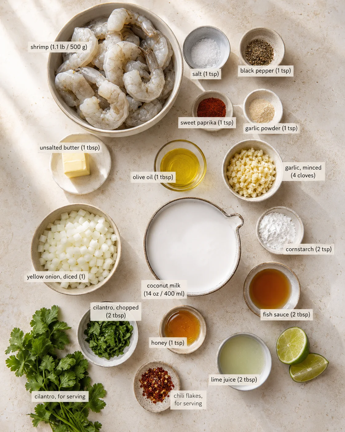 Overhead view of labeled ingredients for a coconut-milk shrimp curry on a beige stone surface.