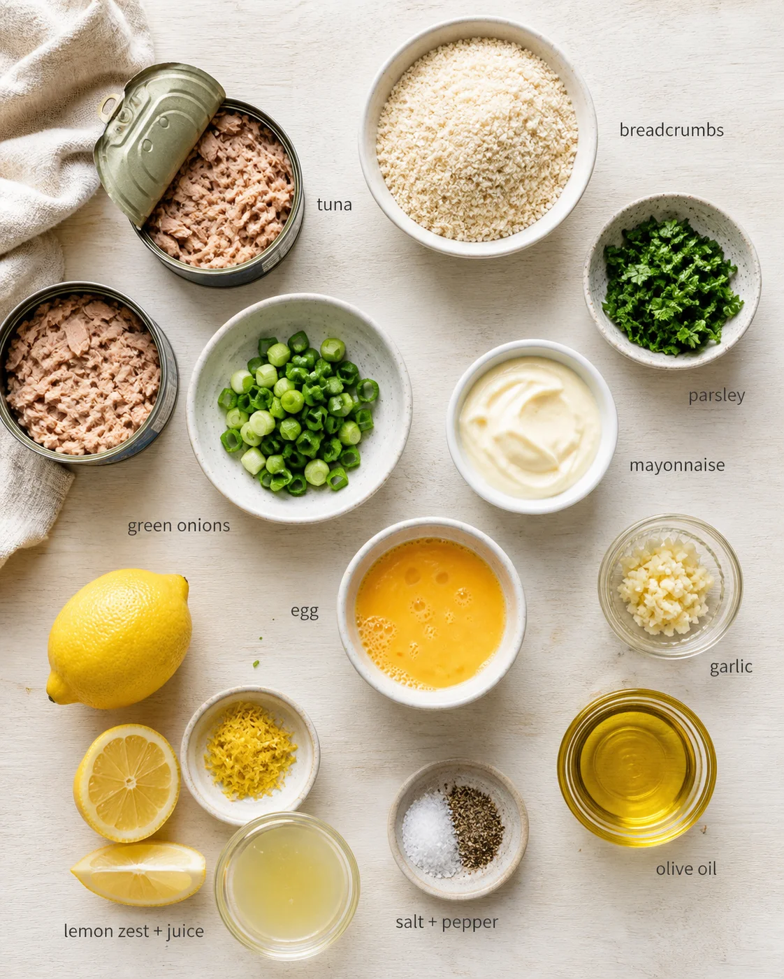 Overhead view of tuna cans, breadcrumbs, green onions, parsley, mayo, lemon, garlic and oils on a light wood surface.