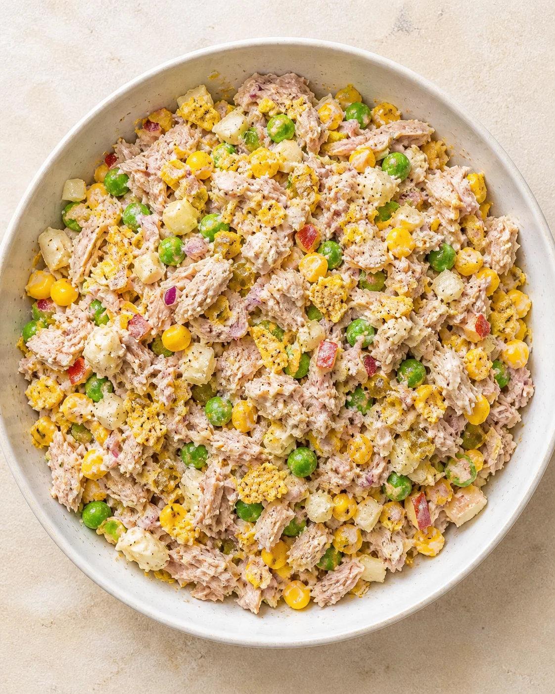 overhead view of a round white bowl filled with tuna salad containing green peas, corn, potatoes, and red onions.