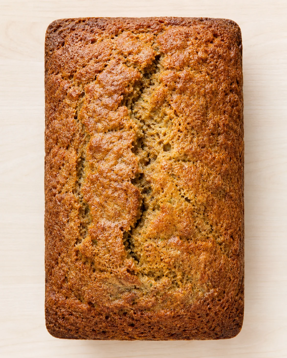 Golden brown banana bread loaf with crackled top on light wooden surface.