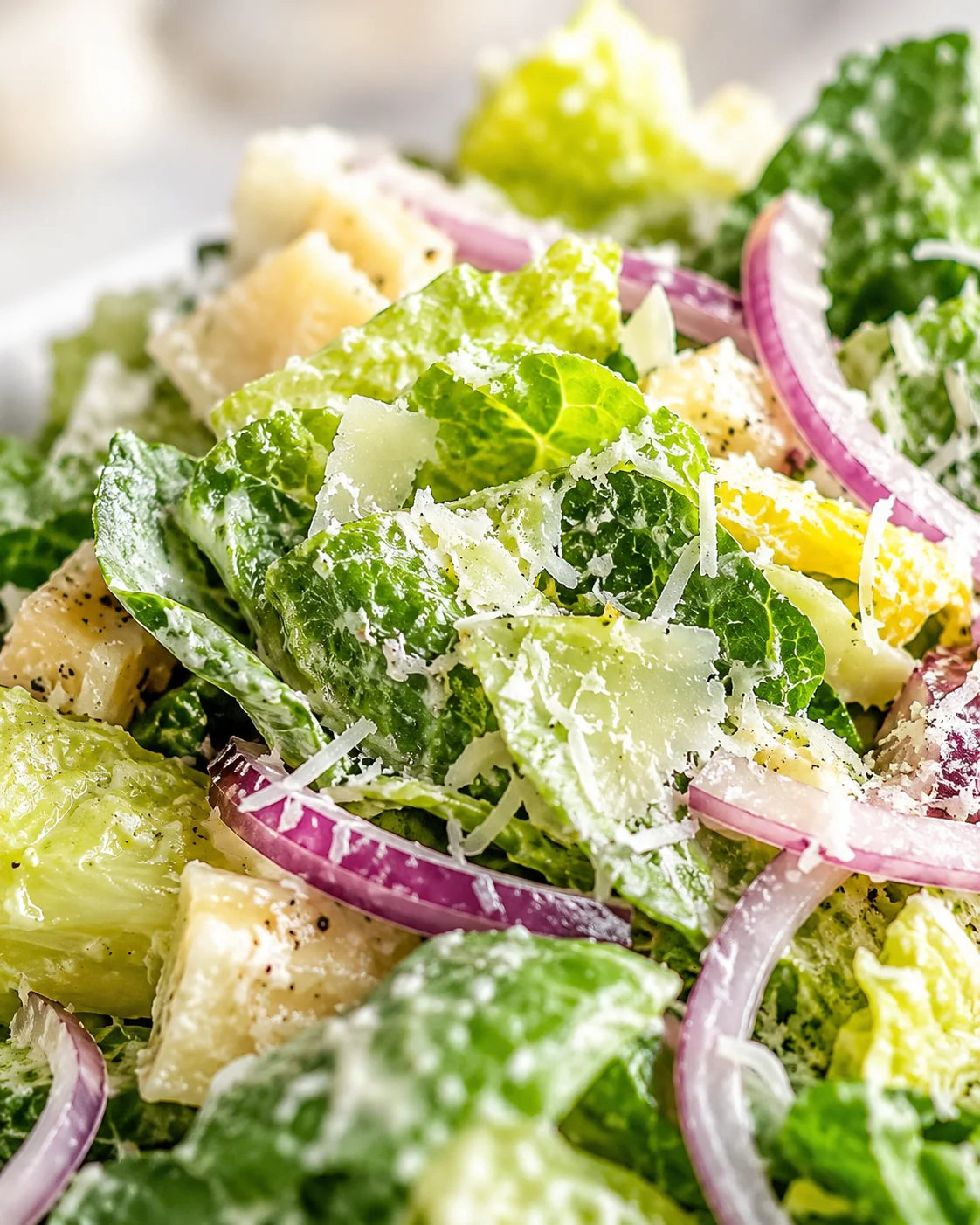 Close-up of crisp romaine lettuce, parmesan shavings, red onion rings, and croutons in creamy dressing.